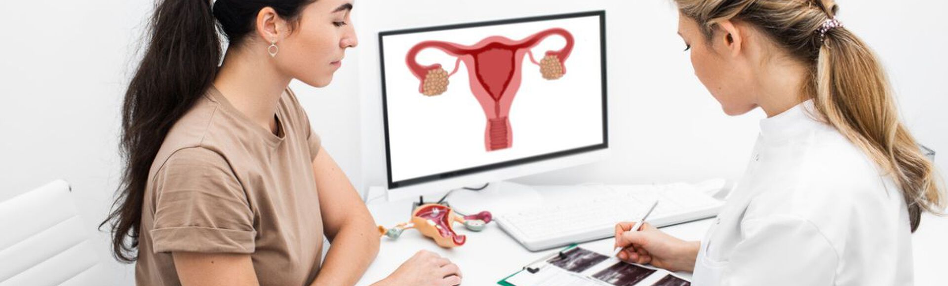 médica explicando um diagnóstico para uma paciente em um consultório. A médica está mostrando um diagrama do sistema reprodutor feminino em um computador