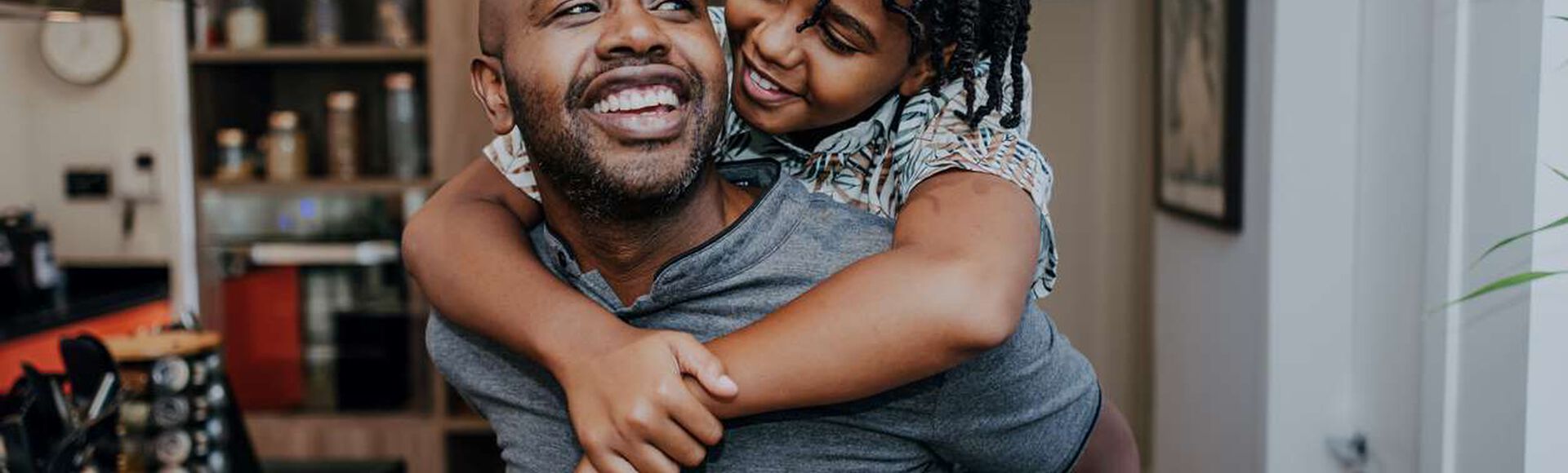 Pai sorrindo enquanto abraça sua filha pequena dentro de casa, representando a paternidade ativa.