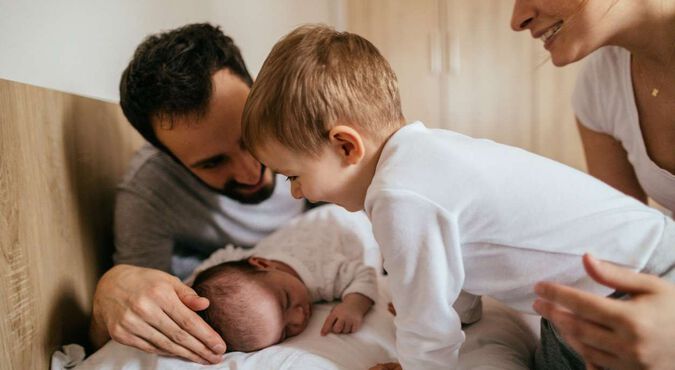 Família na cama, observando um bebê pequeno deitado sobre almofada. O irmão observa o bebê com carinho, o pai e a mãe protegem os filhos.