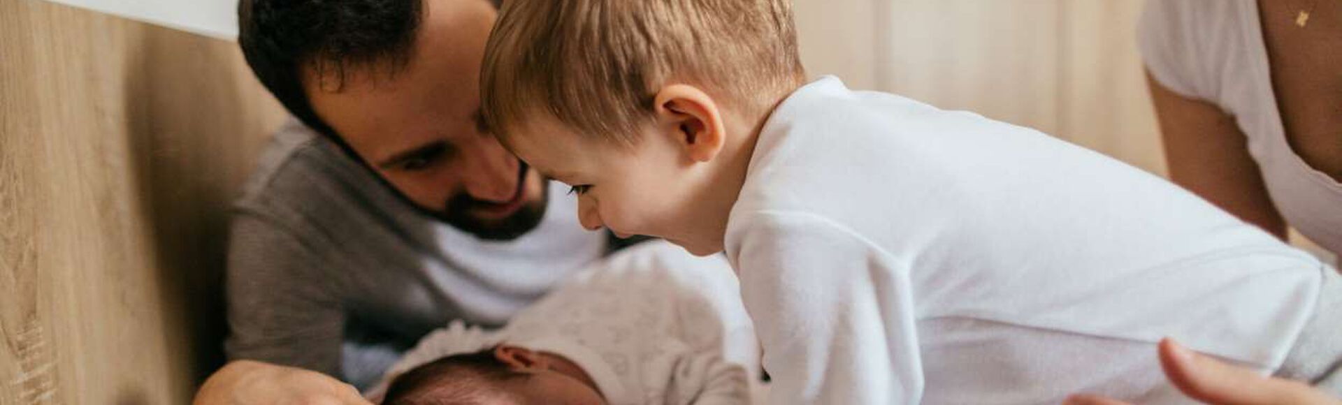 Família na cama, observando um bebê pequeno deitado sobre almofada. O irmão observa o bebê com carinho, o pai e a mãe protegem os filhos.