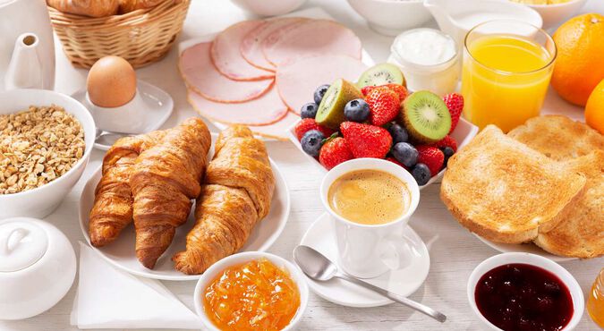Mesa montada com diversas opções de café da manhã: pães, frios, frutas, geleia, suco de laranja, café etc