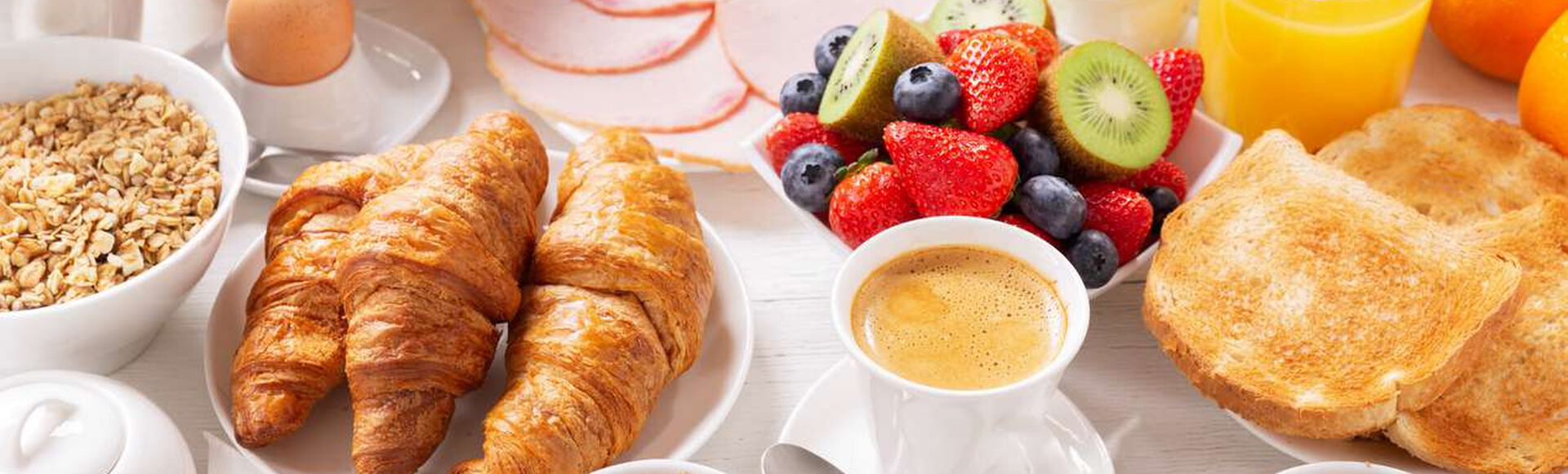 Mesa montada com diversas opções de café da manhã: pães, frios, frutas, geleia, suco de laranja, café etc
