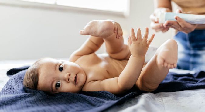Bebê sendo trocado, olhando para a foto, enquanto mulher adulta está preparando seu hidratante.