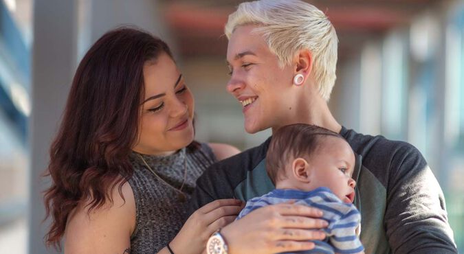 Casal LGBTQIA+ com bebê sorrindo ao ar livre