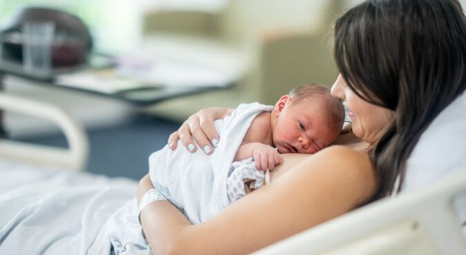 Mamãe e bebê recém-nascido em posição de método canguru. Ambos são brancos e estão deitados na cama do hospital