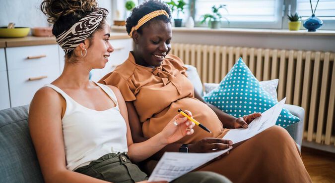 Duas mulheres sentadas no sofá, olhando juntas listas de nomes em papel. Uma delas está grávida.