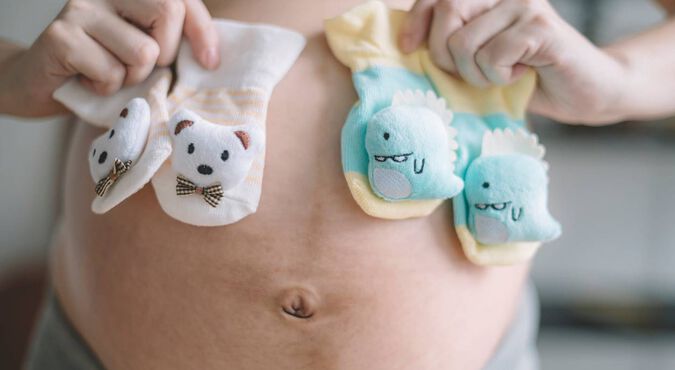 Barriga de uma mulher grávida segurando dois pares de meias de bebê, uma em cada mão.
