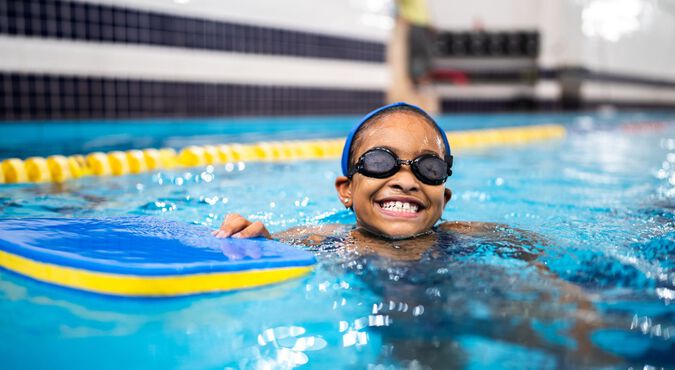 criança sorrindo enquanto nada em uma piscina. A criança está usando óculos de natação e uma touca azul, segurando uma prancha flutuante azul e amarela.