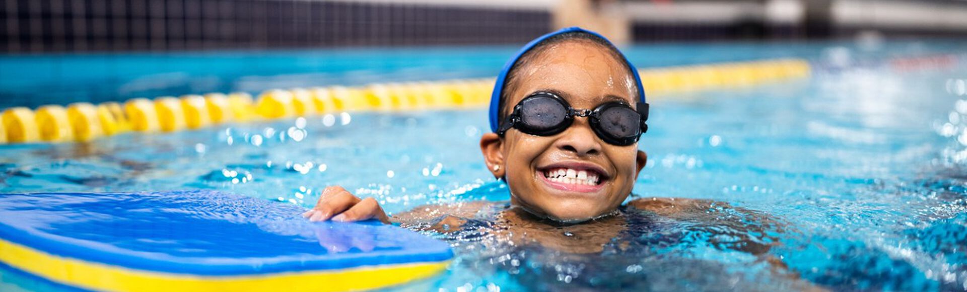 criança sorrindo enquanto nada em uma piscina. A criança está usando óculos de natação e uma touca azul, segurando uma prancha flutuante azul e amarela.