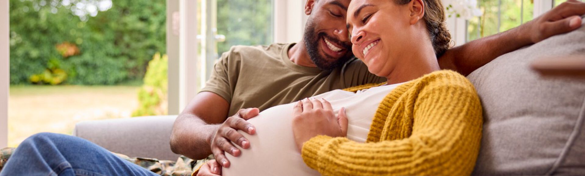 Casal feliz em artigo sobre sintomas de gravidez. Eles são negros e estão no sofá com a mão na barriga da mulher grávida