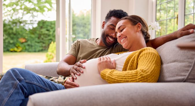 Casal feliz em artigo sobre sintomas de gravidez. Eles são negros e estão no sofá com a mão na barriga da mulher grávida