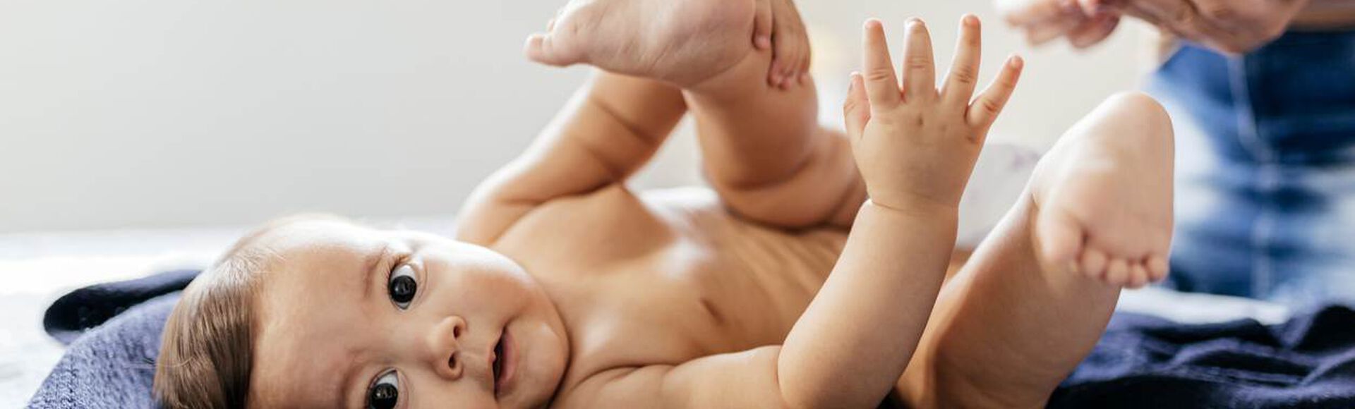 Bebê olhando para a câmera, no momento da troca de fralda. Na foto, vemos sua mãe aplicando pomada contra assaduras.