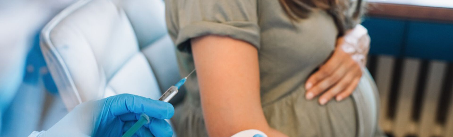 Mulher branca gestante recebendo vacina no consultório médico. Rosto da grávida e de quem aplica a vacina não aparecem.
