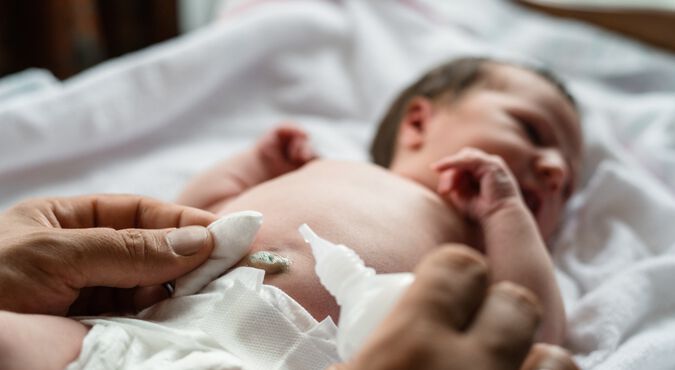 Recém-nascido branco, deitado no trocador, tem resquício de cordão umbilical limpo. Aparece apenas a mão da pessoa que limpa