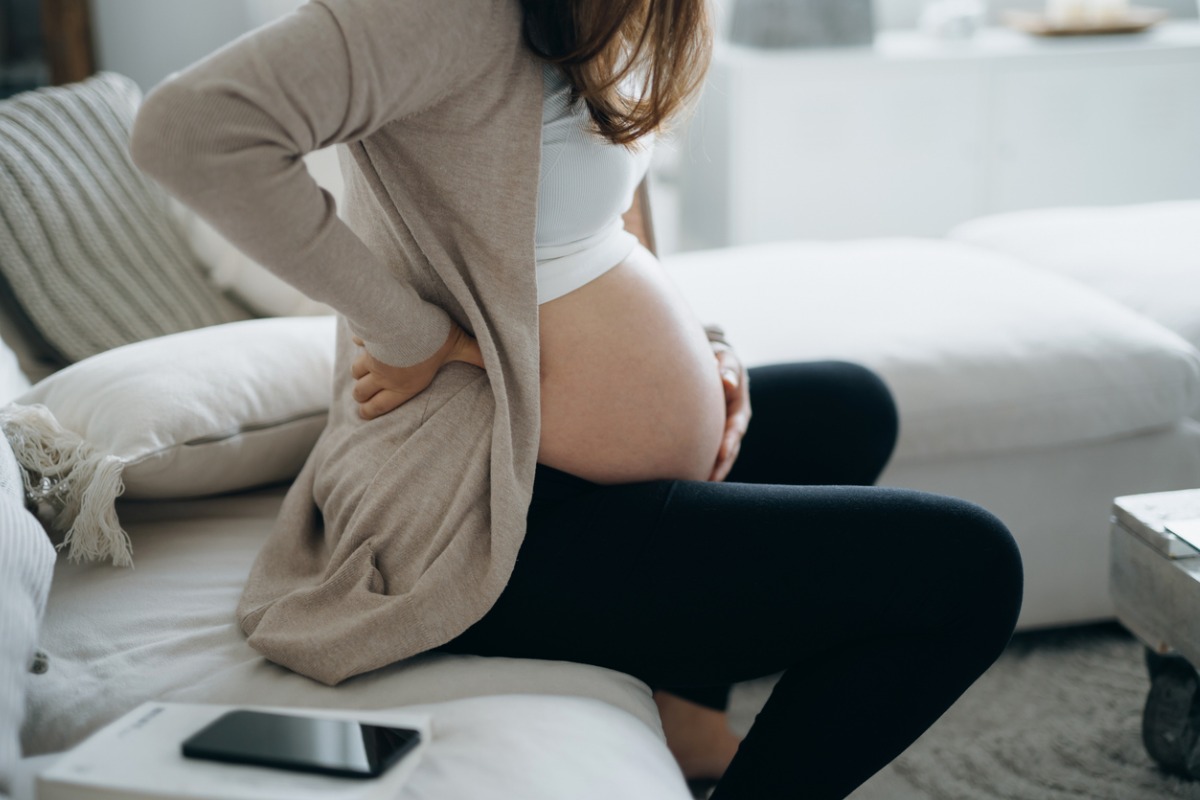 Mulher branca sentada na beira do sofá com a mão na barriga em artigo sobre primeiros sintomas de gravidez. Rosto não aparece 