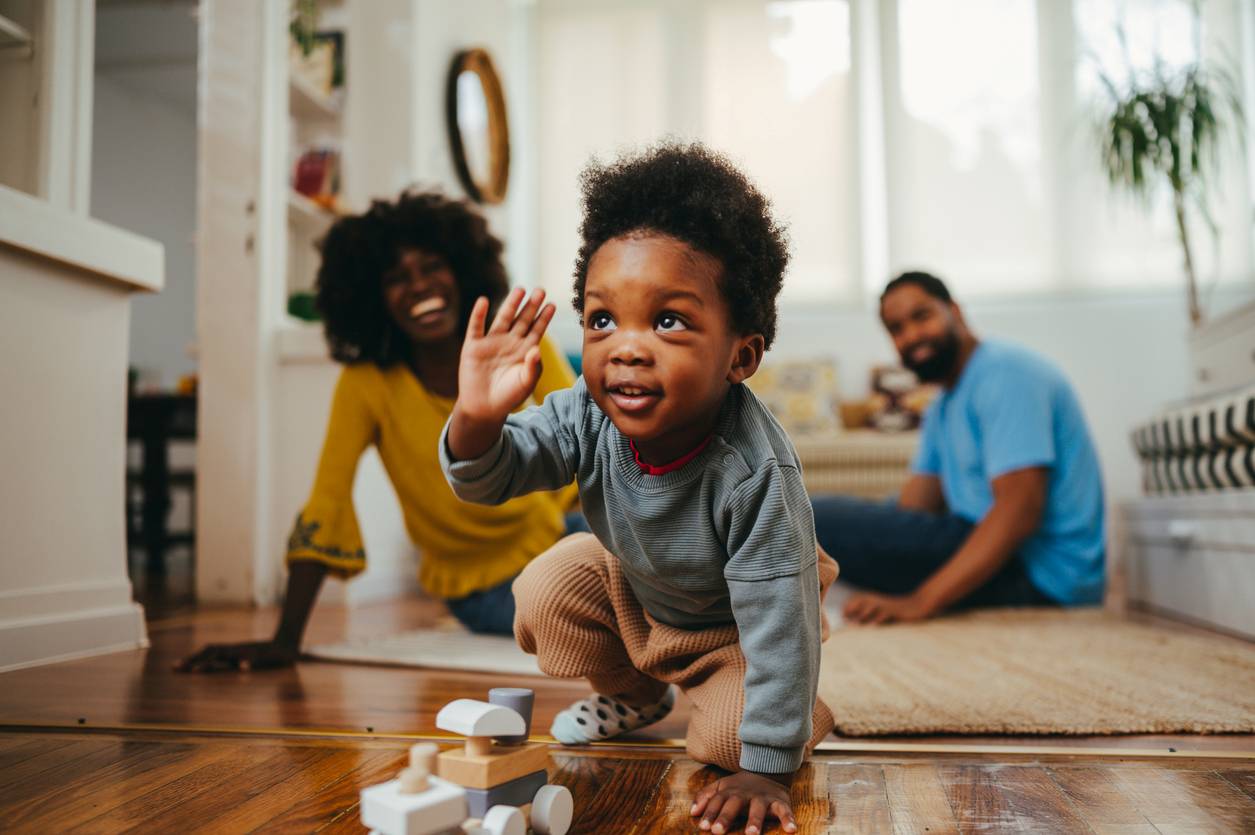 Bebê engatinhando, seus pais sentados atrás dele na sala de estar. Todos com cabelos crespos de tipos diferentes.