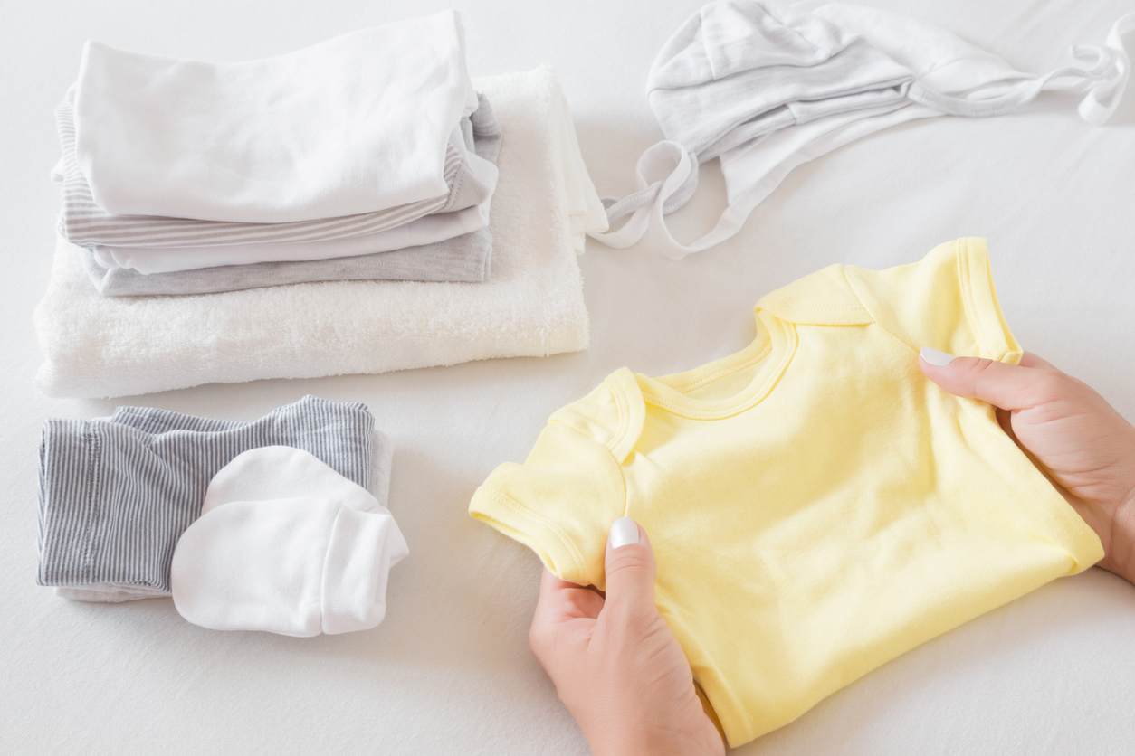 Detalhe de mãos segurando camisetinha amarela ao lado de pequenas pilhas de roupa de nenê dobradas.