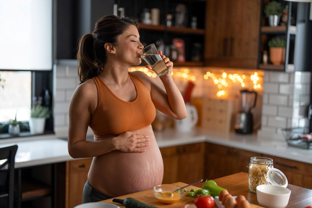 Mulher grávida está bebendo água em sua cozinha enquanto acaricia a barriga