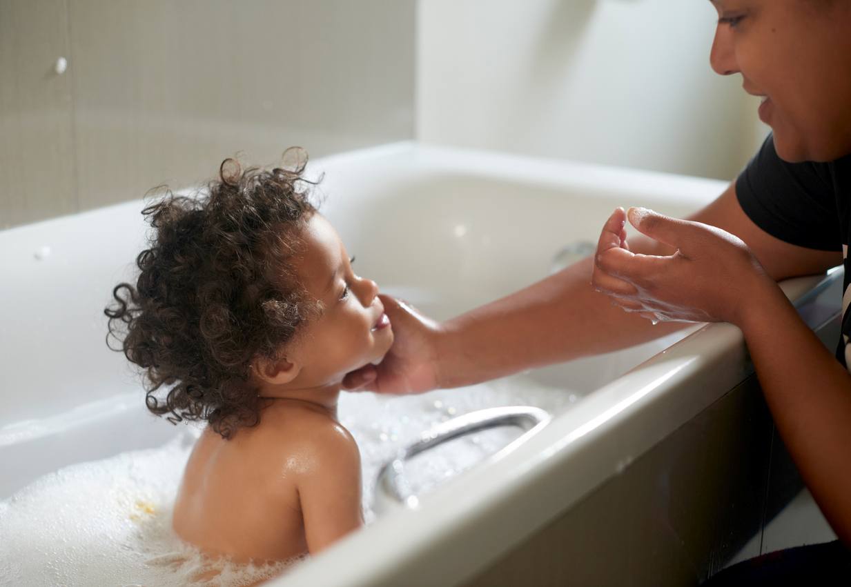 Mãe acariciando o rosto de seu bebê que está sentado na banheira, com os cabelinhos todos arrepiados.