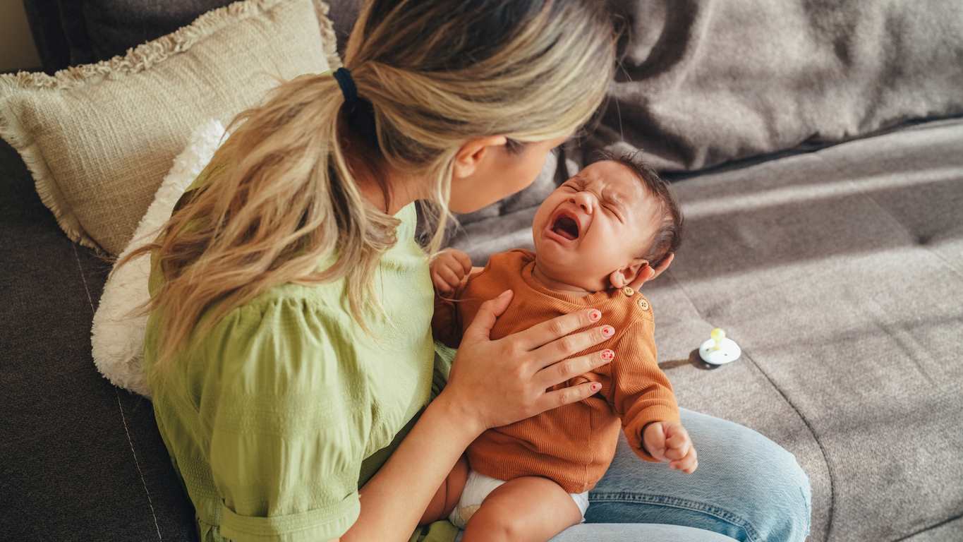 Sentada no sofá, mamãe conforta bebê de colo chorando muito. Os dois são brancos, a mãe está de verde e o bebê, de marrom