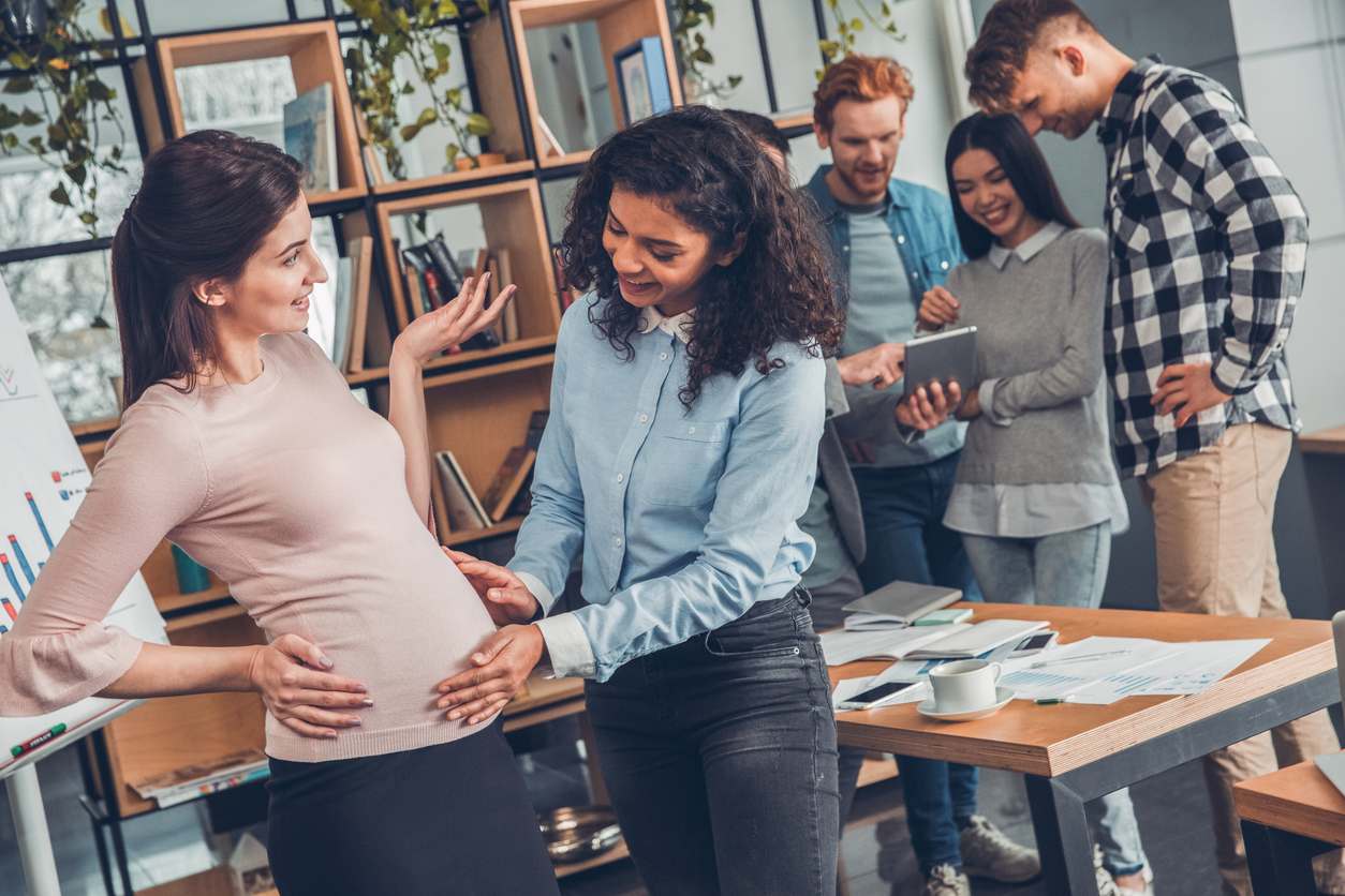 Colegas se divertem no café da empresa, enquanto gestante conversa com uma mulher que toca sua barriga.