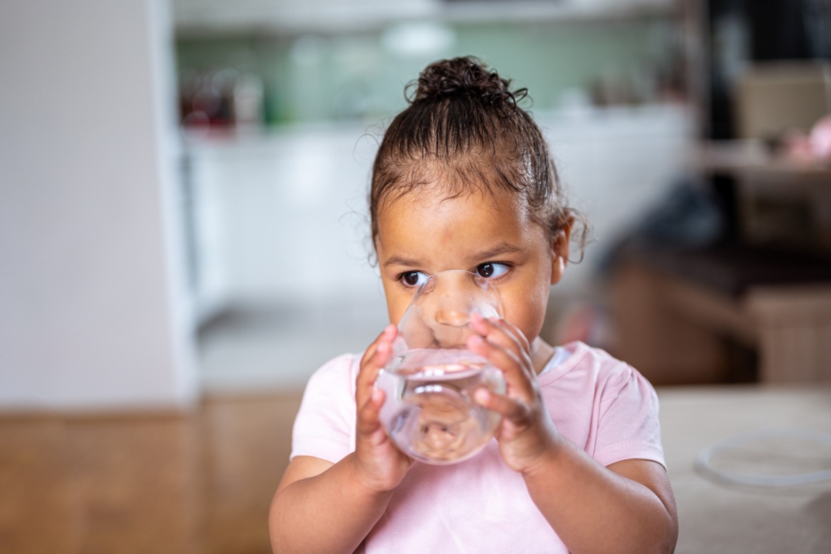 Criança pequena mestiça toma água no copo para evitar infecção urinária infantil. Ela veste camiseta rosa e fundo está sem foco