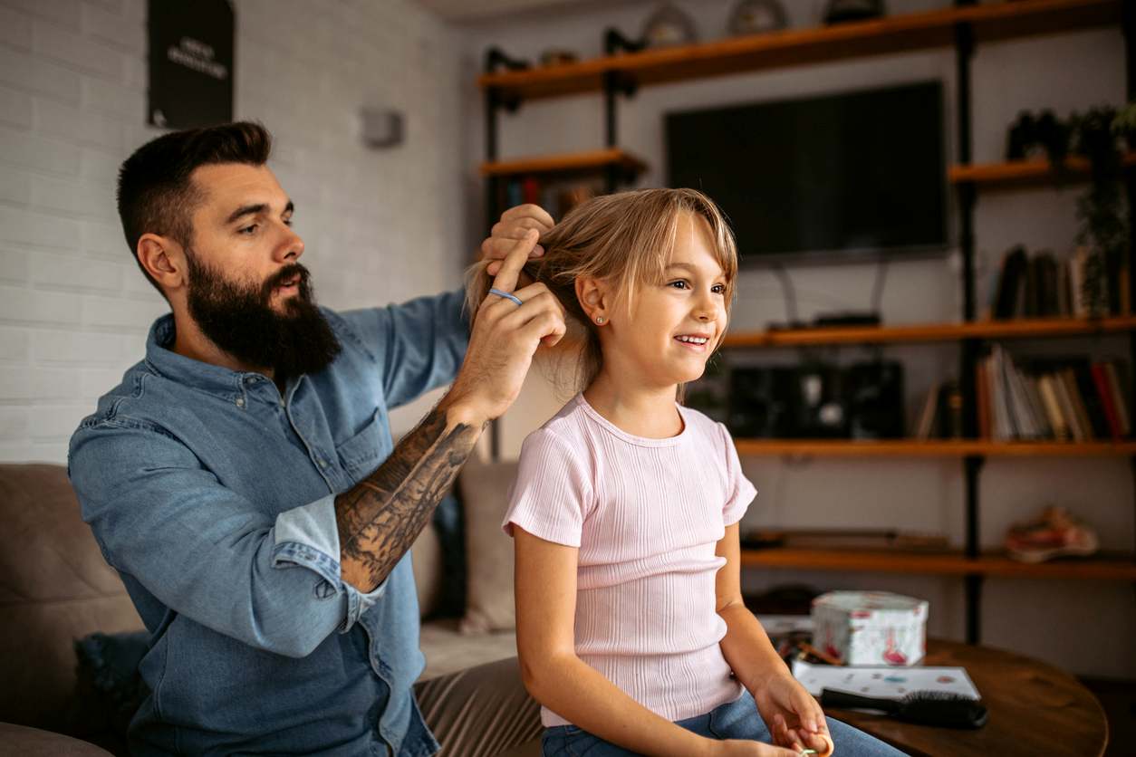 Menina de cabelo liso, sentada em uma mesa de centro enquanto o pai cuida de seus cabelos.