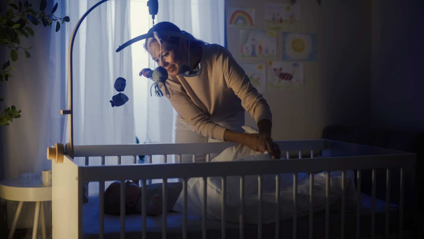 Com luz noturna ambiente, mamãe checa seu bebê que dorme no bercinho.