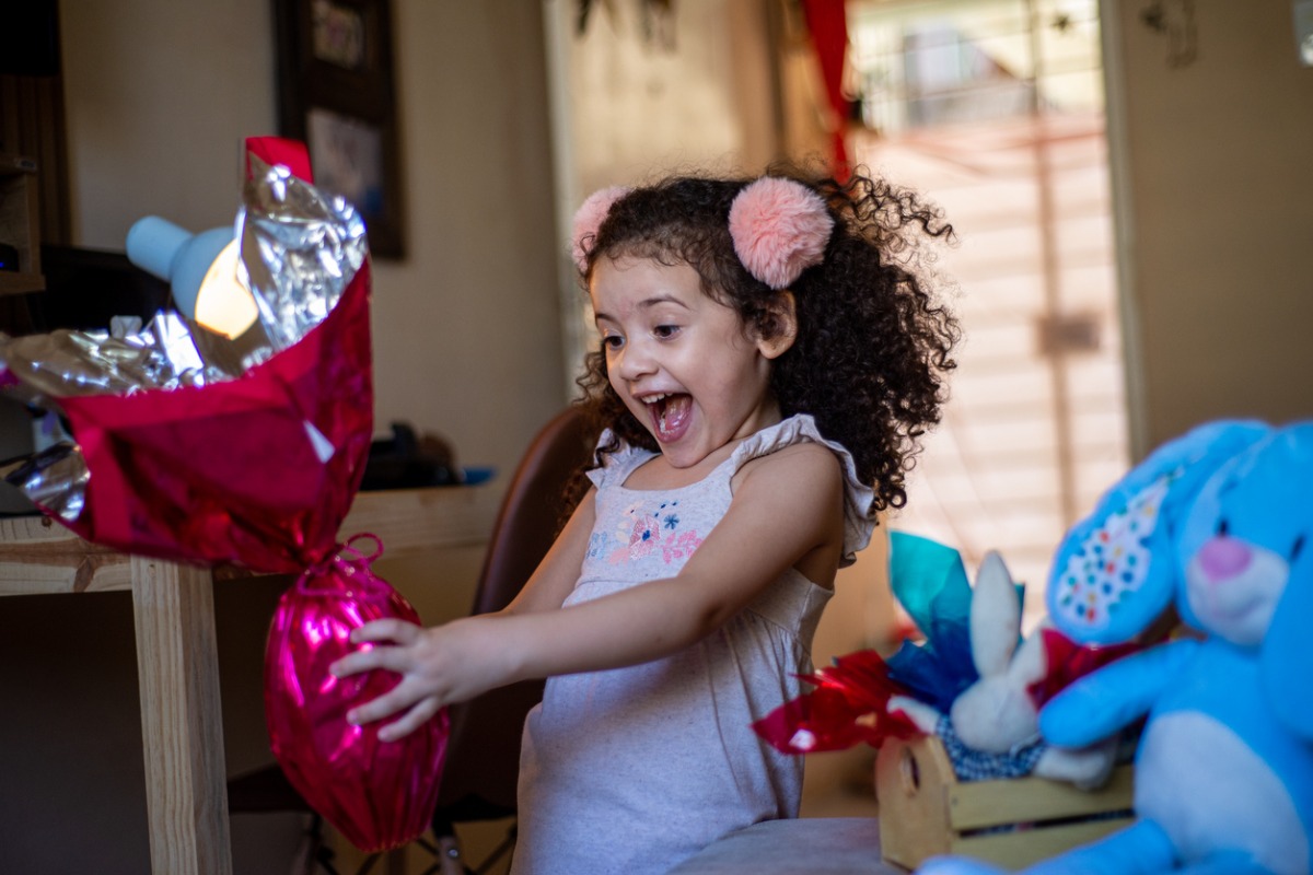 Menina de cabelo cacheado escuro muito feliz com seu ovo de Páscoa. Ao seu lado há um coelho de pelúcia e uma cesta de Páscoa 