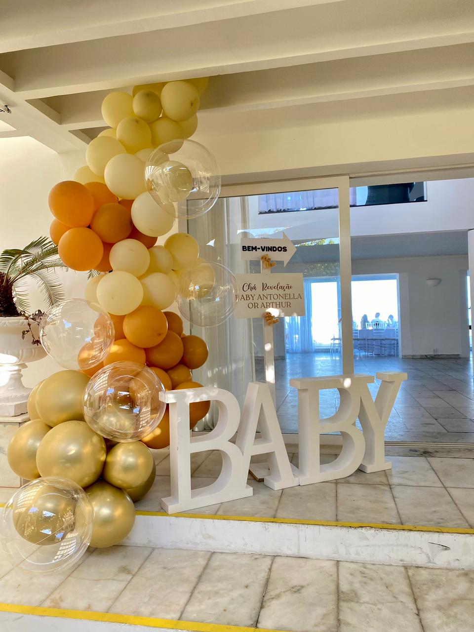 Entrada de festa de chá revelação com balões amarelos, dourados e alaranjados, com letras formando a palavra bebê em inglês 