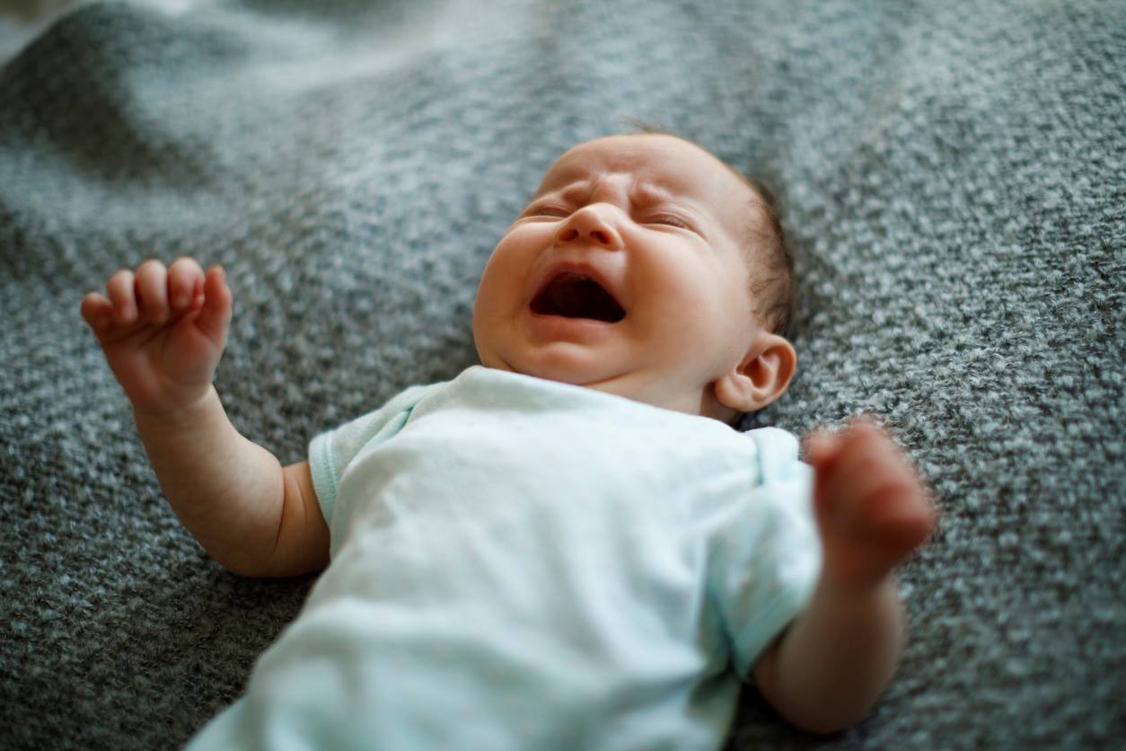 Bebê chorando muito irritado durante a hora da bruxa. Ele é branco, está de roupinha verde claro e deitado sobre coberta verde 