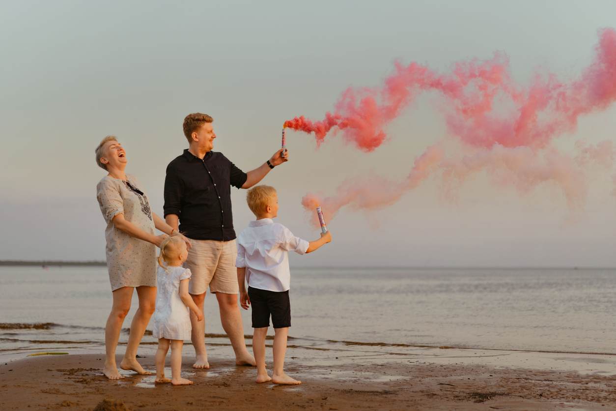 Família comemorando revelação de gênero com fumaça colorida em praia.
