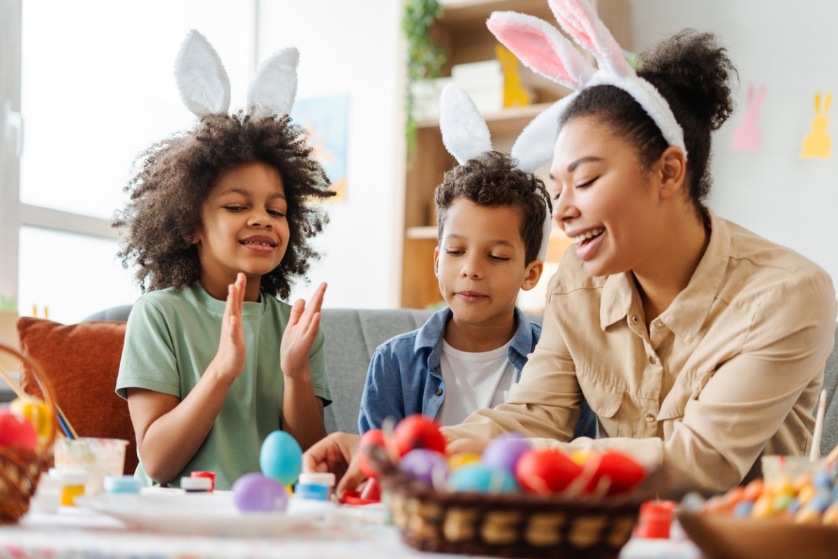 Mulher mestiça adulta com dois meninos pintando ovos de Páscoa. Na mesa, ovos coloridos e a família usa orelhas de coelho 