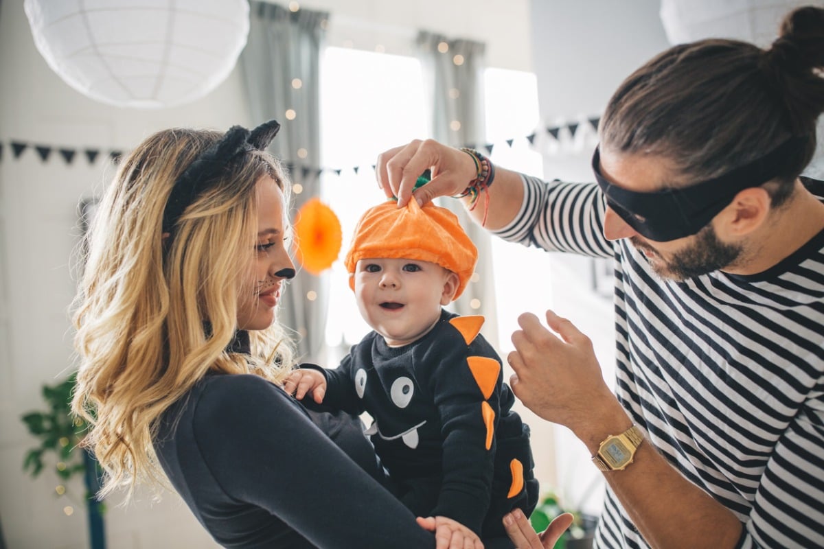 Mãe com bebê no colo enquanto o pai ajusta o chapéu da fantasia do pequeno antes de ir para o bloquinho de carnaval