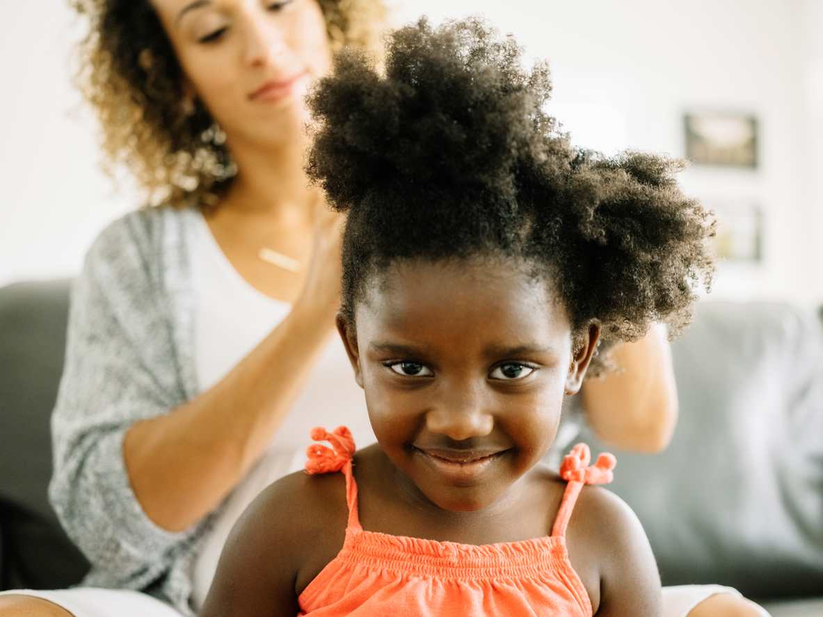 Menina de cabelo crespo, olhando para a câmera enquanto sua mãe cuida de seus cabelos.