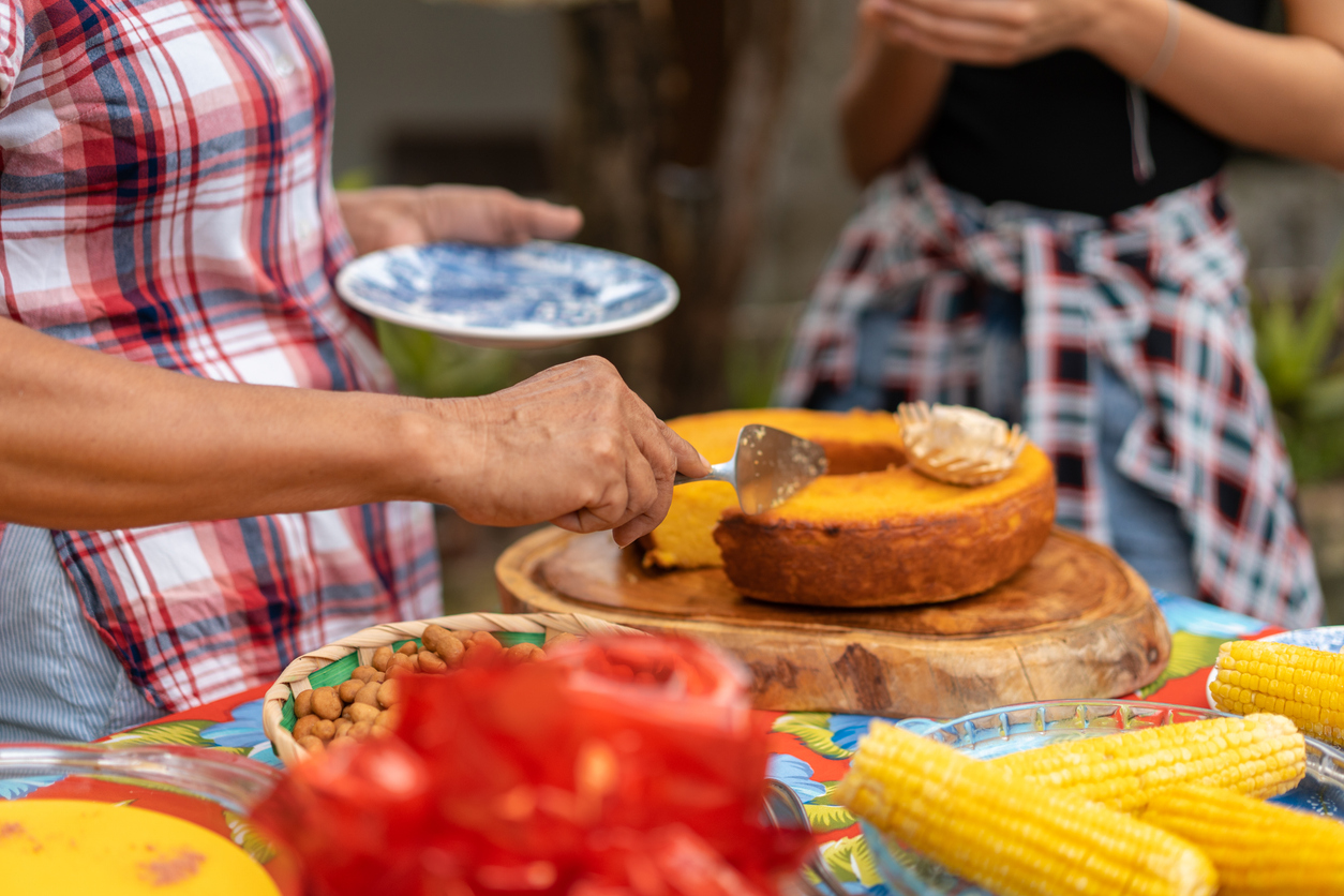 Mesa com quitutes para charraiá de fraldas. Bolo de fubá, milho cozido, amendoim e outros. Há duas pessoas ao redor mesa. 