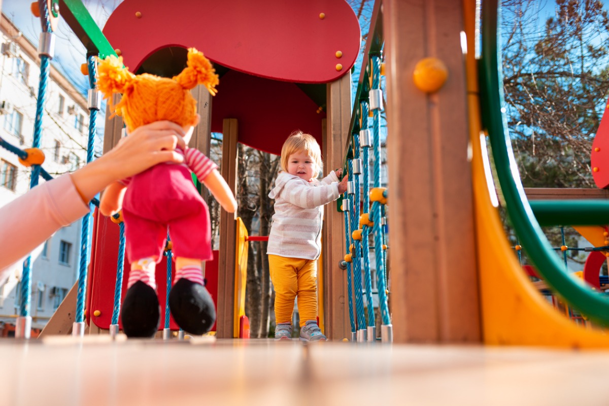 Criança pequena brincando no parque em artigo sobre sinais de autismo em bebês. Uma pessoa aponta uma boneca para a bebê 