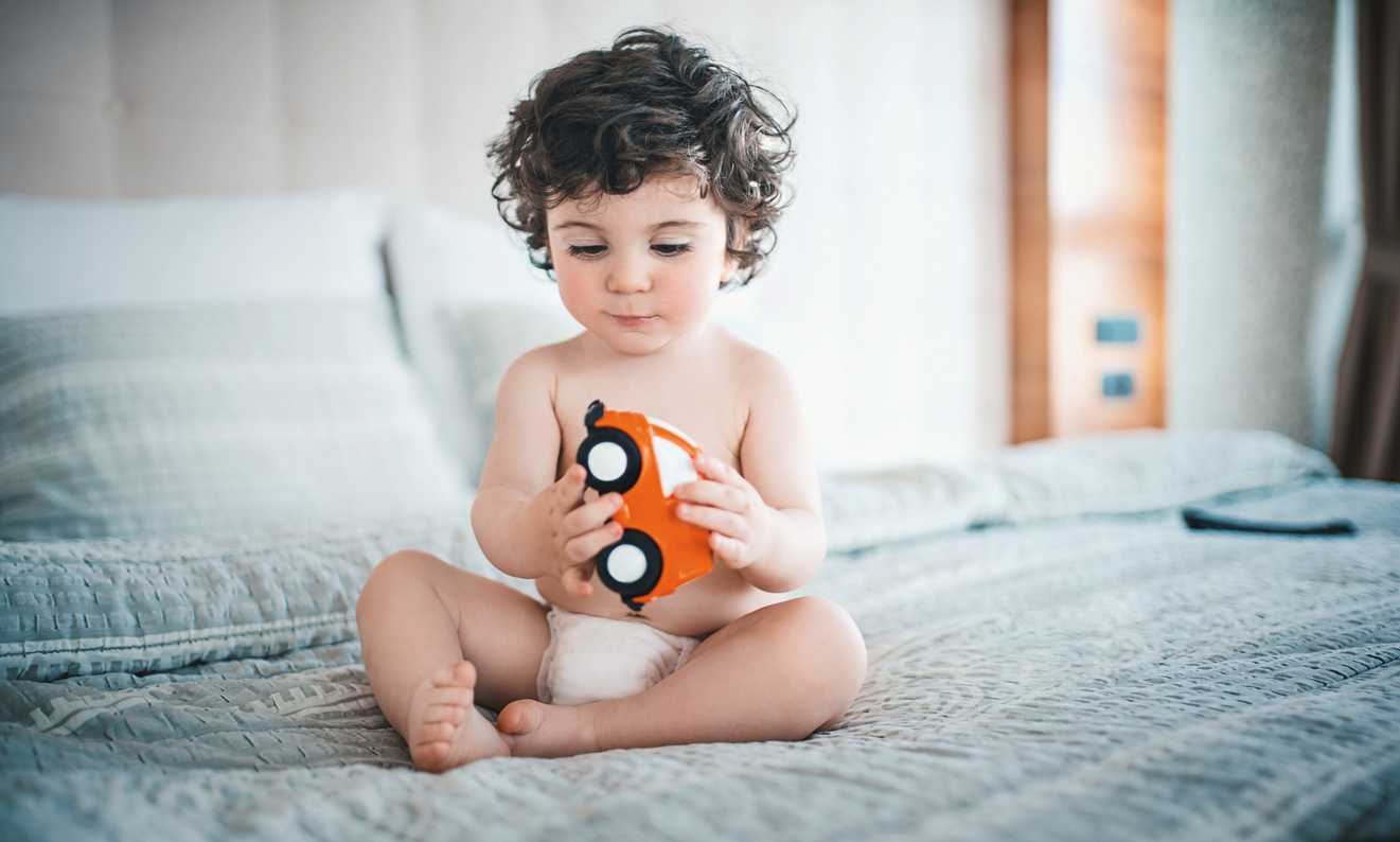 Bebê sentado na cama, de fraldinha, brincando com carrinho. O charme é seu cabelo cacheado.