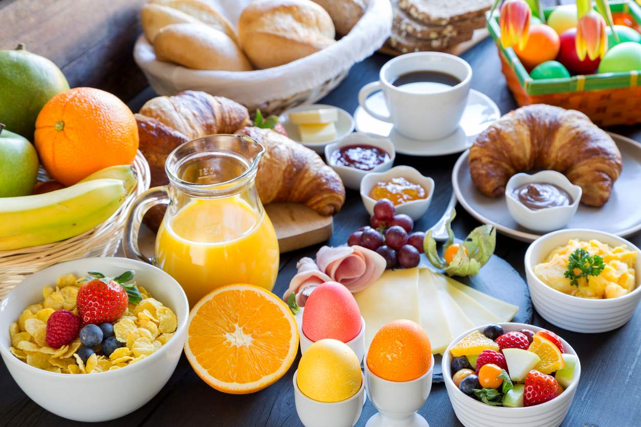 Mesa com delicioso brunch servido. Na imagem vemos ovos, frutas, croissants, pães, suco de laranja, cereal, frios e muito mais.