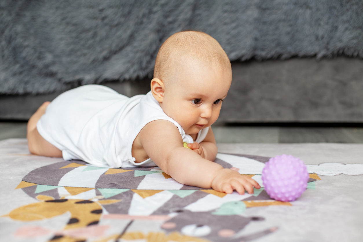 Bebê de bruços sobre tapete de bichinhos, na sala, tentando pegar uma bolinha lilás.